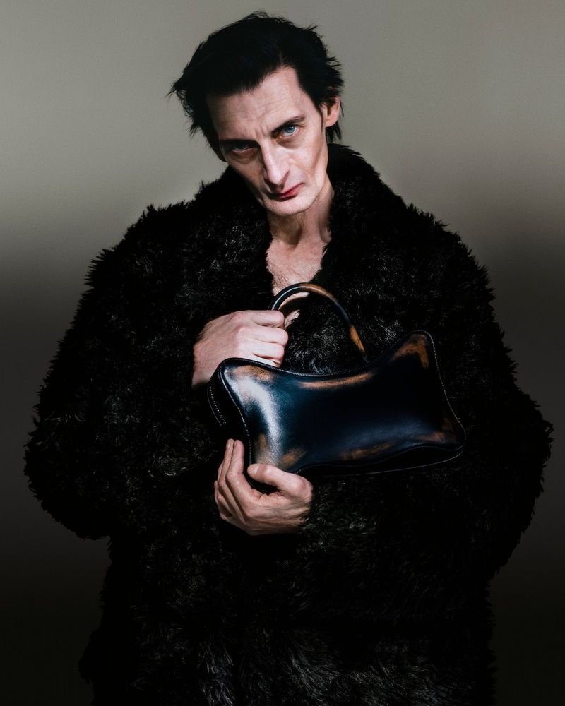 Adult male in a black furry coat holds a dark, shiny purse in a studio setting with a muted background and soft lighting.