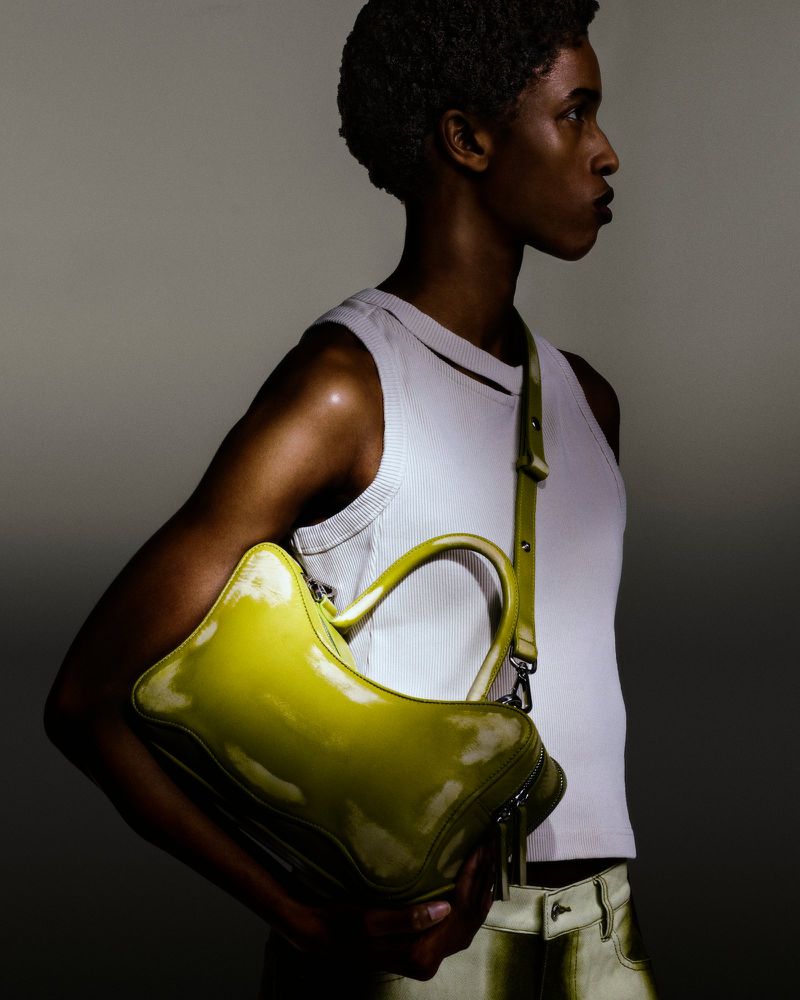 Model presents a glossy chartreuse-colored handbag with a shoulder strap in a studio shot against a soft grey backdrop.
