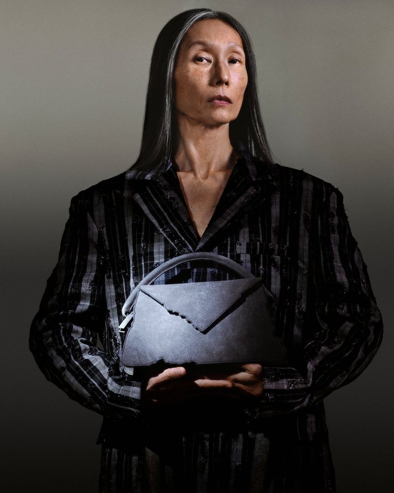 Elegant older woman in a pinstripe suit holds a gray envelope-style handbag in a studio setting with soft lighting, creating a sophisticated fashion portrait.