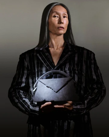 Elegant older woman in a pinstripe suit holds a gray envelope-style handbag in a studio setting with soft lighting, creating a sophisticated fashion portrait.