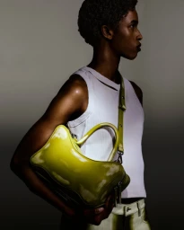 Model in profile wearing a white ribbed tank top holding a glossy chartreuse handbag against a dark gradient studio background.
