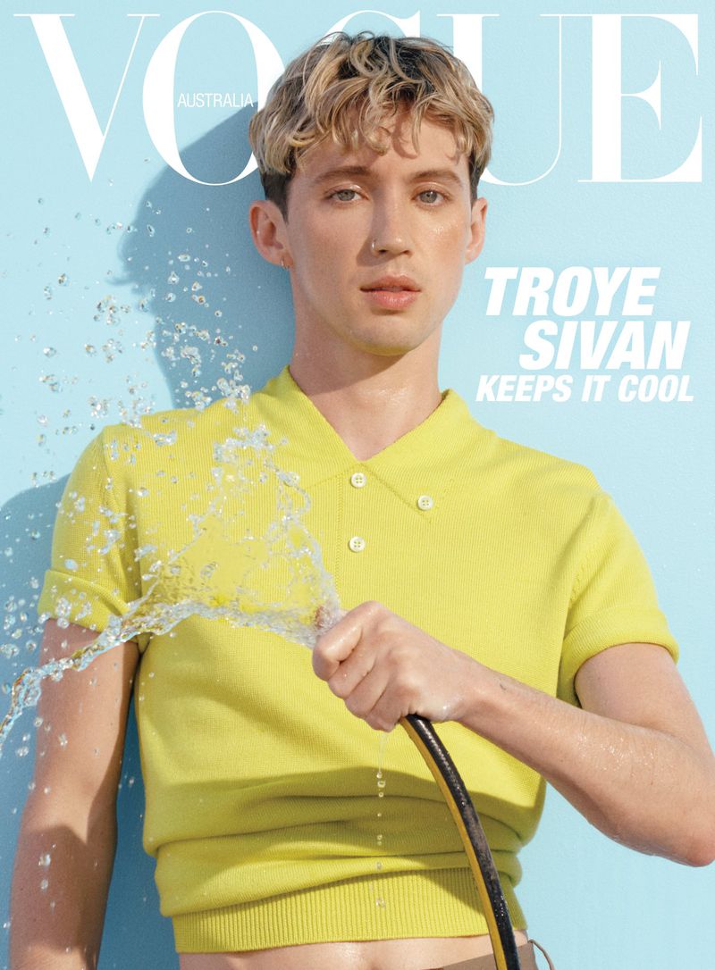 Troye Sivan on magazine cover, blonde curly hair, yellow collared shirt, holding water hose against light blue studio background.