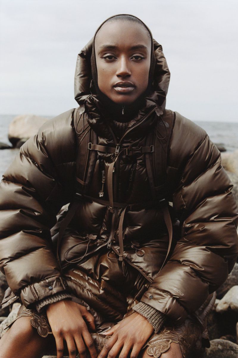 A dark-skinned female fashion model wearing a brown puffer coat and skirt poses outdoors near a rocky ocean shoreline in a medium shot.