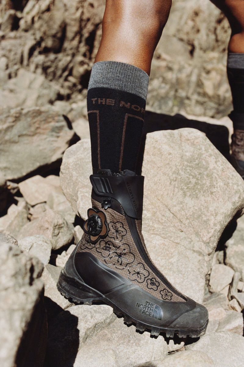 Close-up of a black and tan The North Face hiking boot with floral design on a rocky terrain, paired with gray and black socks.