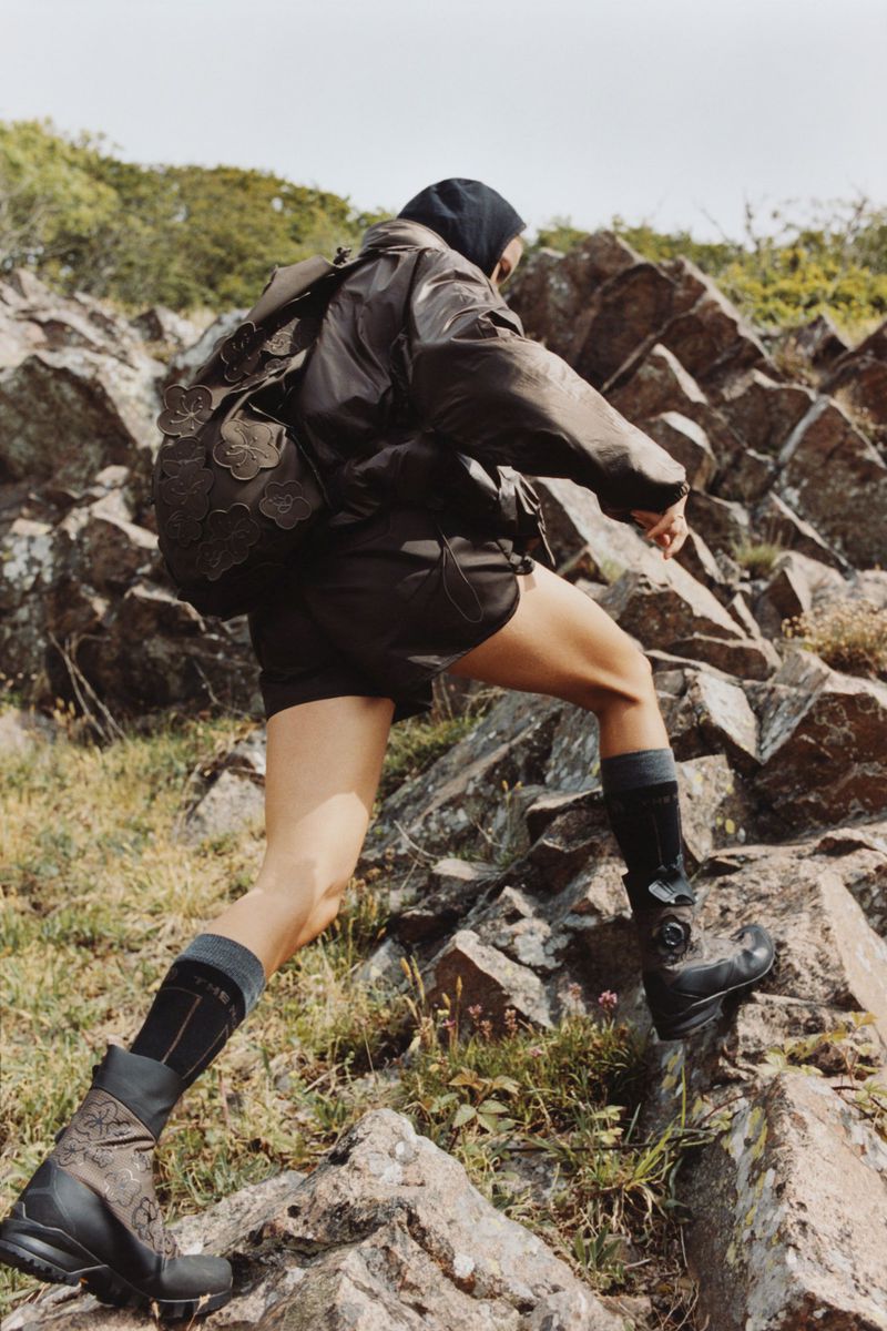 Athletic woman with backpack hikes on rocky terrain, wearing shorts, long socks, and patterned black boots in an outdoor setting.
