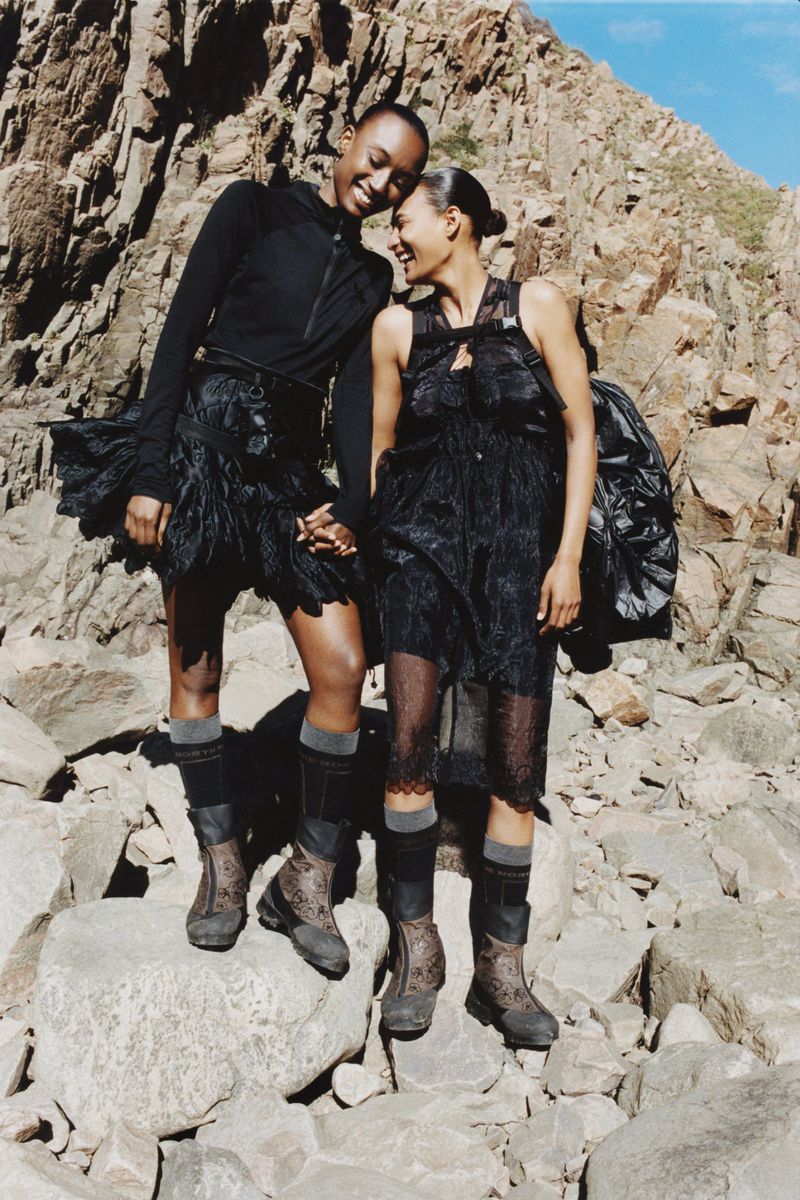 Two women in black dresses and boots pose on a rocky mountain terrain with a bright sky, showcasing outdoor fashion and a rugged landscape.