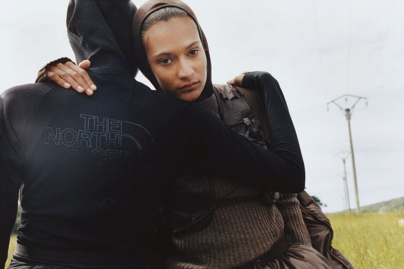 Two people embrace in an outdoor setting; one wears The North Face hoodie. A landscape includes grassy fields and utility poles.