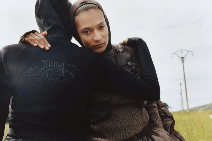 Two people embrace in an outdoor setting; one wears The North Face hoodie. A landscape includes grassy fields and utility poles.