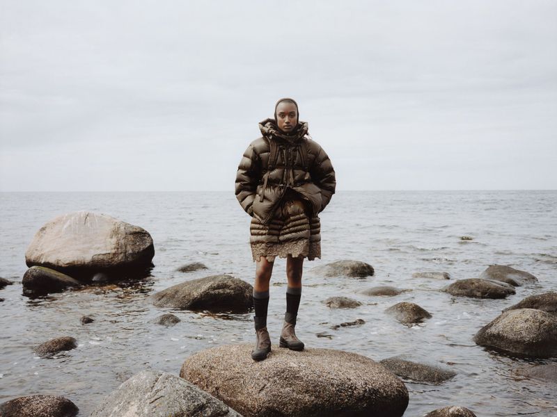 Woman in a puffy coat standing on a rock in the water on a cloudy day. Fashion shot in a natural, rocky environment. 