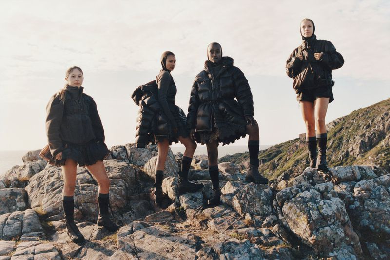 Four women model black outerwear and boots, standing on a rocky outcrop against a bright sky and distant green landscape in a fashion editorial.