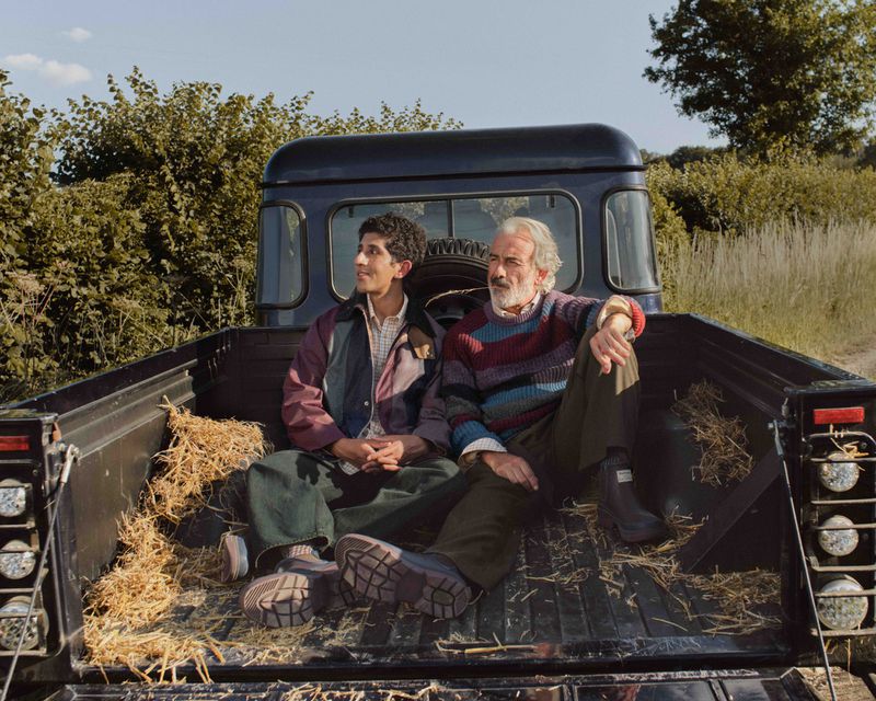 Two men of mixed ethnicity sit in the back of a pickup truck in a rural setting. The truck bed is lined with straw.