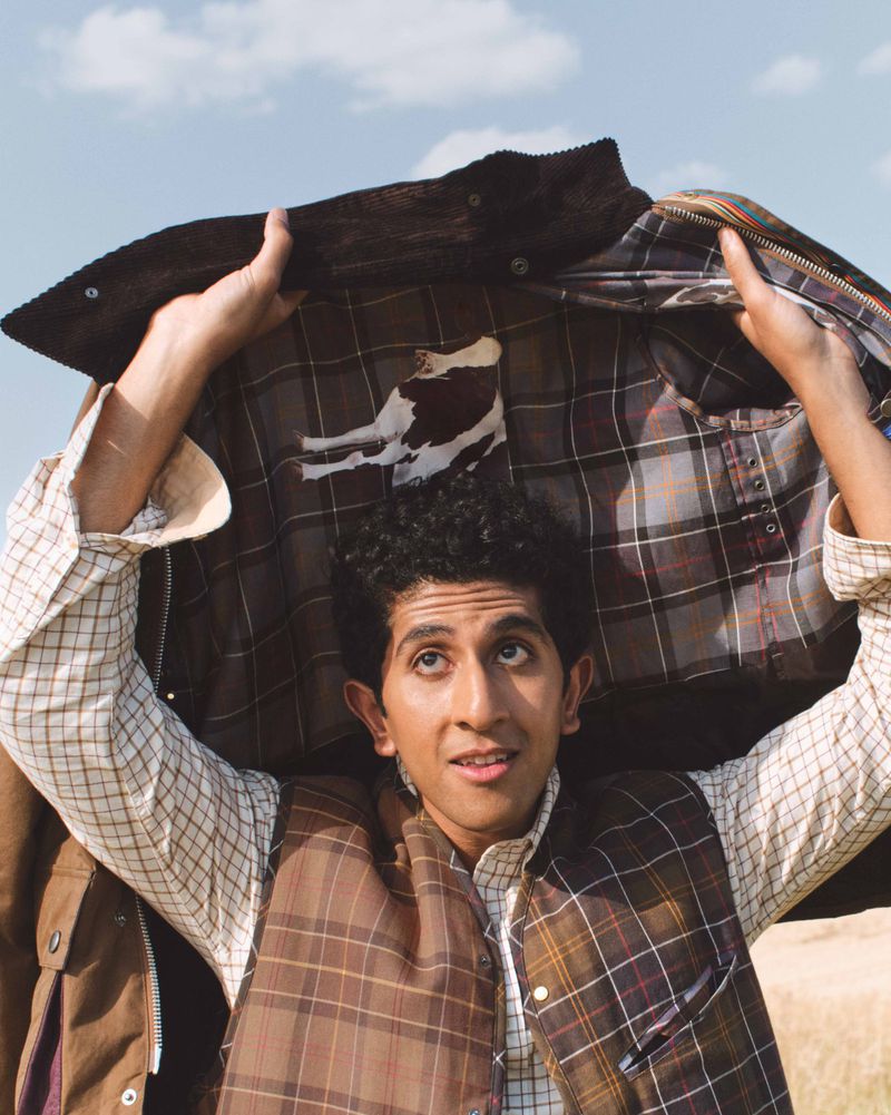 Adult male smiles while holding a brown plaid jacket over his head outdoors with a blue sky background, showcasing fashion style.
