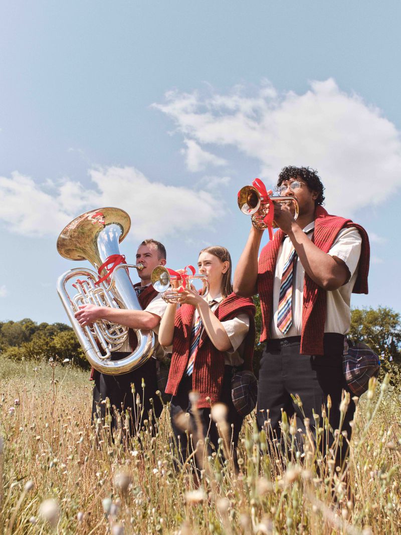 Outdoor image of a musical band composed of three people playing various brass instruments on a field under a cloudy sky.