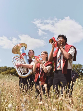 Outdoor image of a musical band composed of three people playing various brass instruments on a field under a cloudy sky.