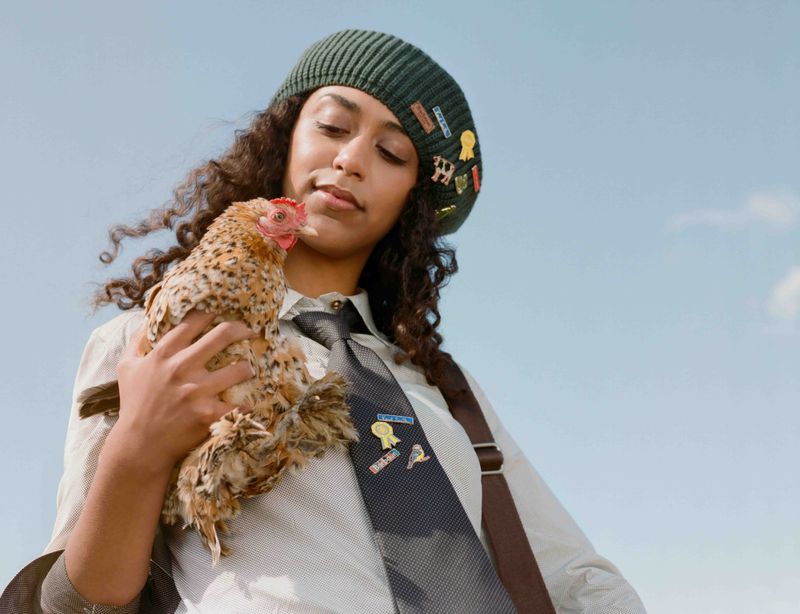 Woman in beret holds chicken outdoors against blue sky. The bird has light brown speckled plumage and she wears a collared shirt with tie.