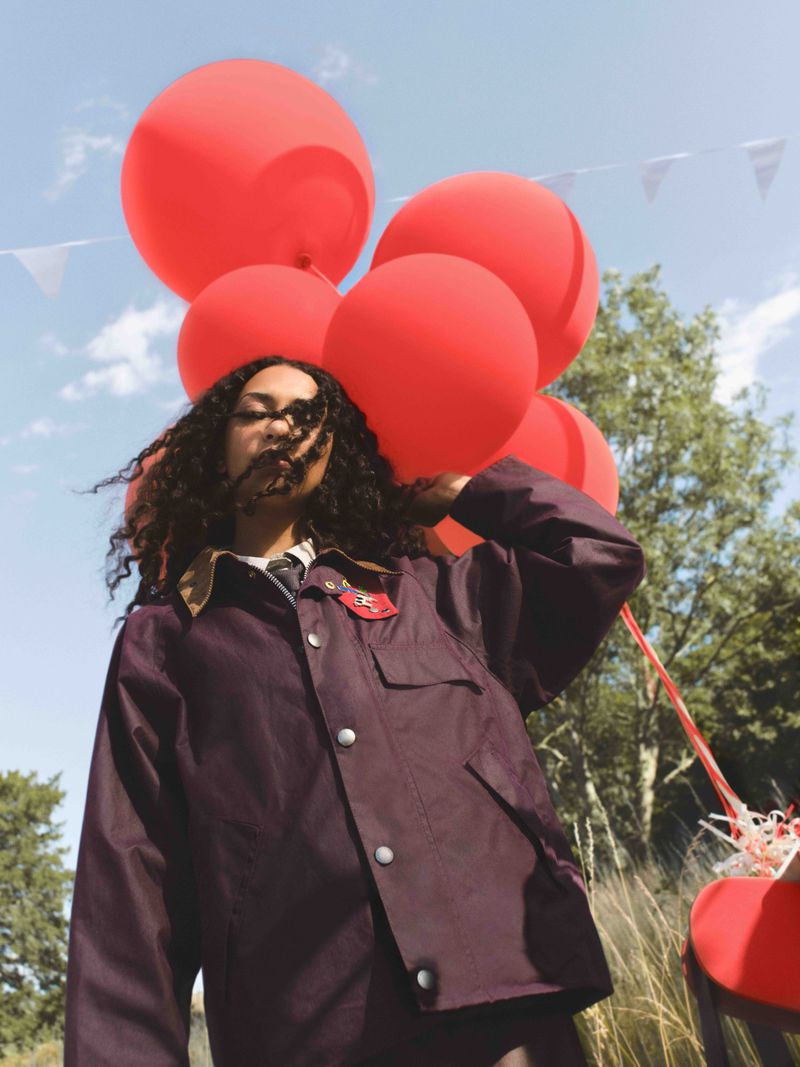 A woman in a brown jacket stands outdoors holding red balloons against a blue sky backdrop with trees and distant white flags.