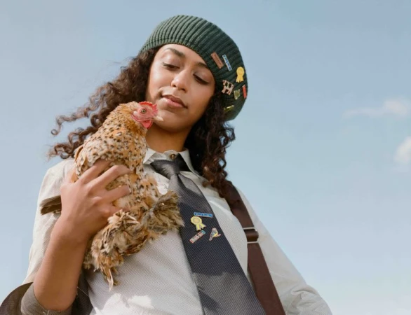Woman wearing a dark green beret and collared shirt with a necktie holding a small speckled chicken against an open sky.