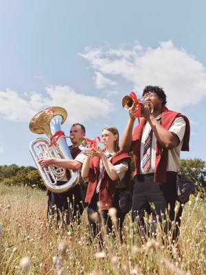 Three brass band musicians wearing burgundy knitwear and striped ties play instruments in a sunlit grassy field.