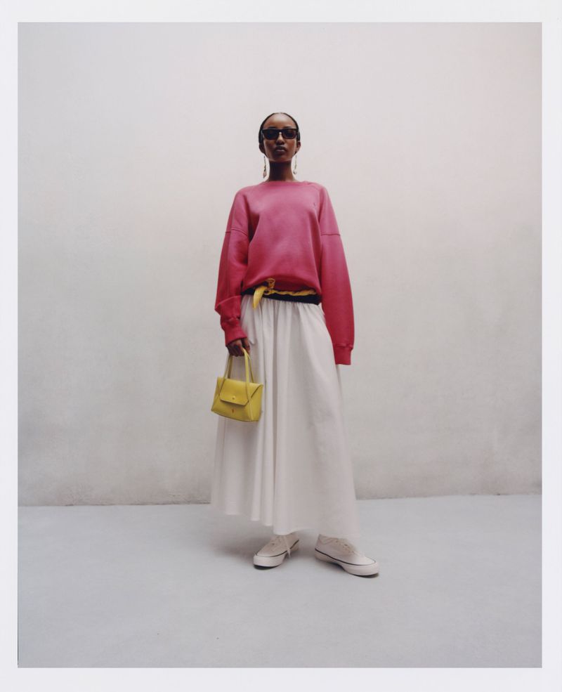Woman in pink sweatshirt, white skirt, sneakers, and sunglasses holding a yellow purse, posing indoors against a textured wall.