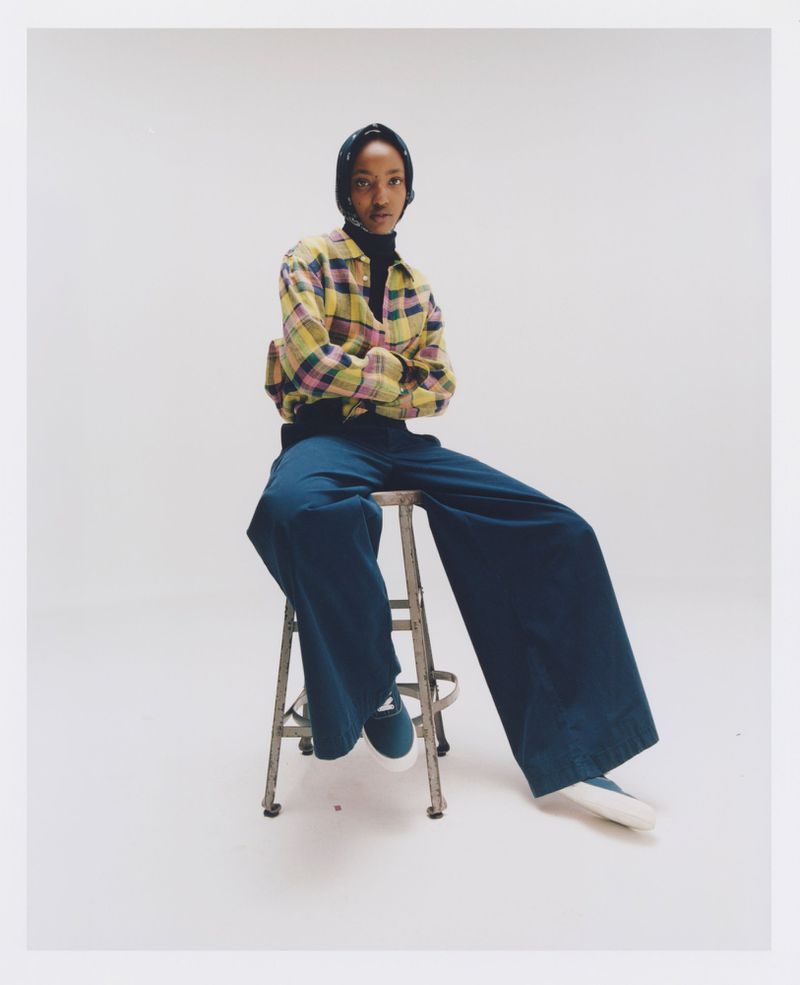 A slender person with arms crossed poses in a yellow plaid shirt and blue pants, seated on a stool, against a bright white backdrop.