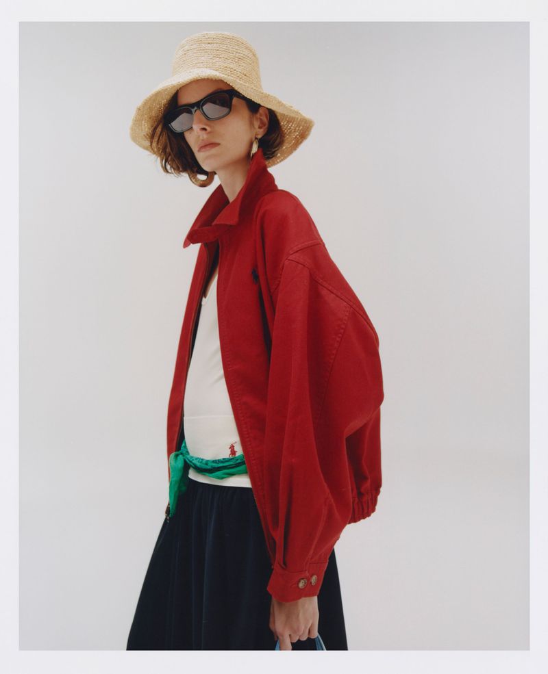 A fashionable woman with a straw hat and sunglasses poses in a red jacket, white top, and navy skirt against a white background.