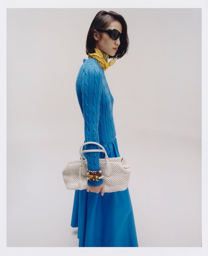 Stylish woman in a blue dress and sunglasses carries a white woven handbag against a bright backdrop, showcasing a fashion-forward look.