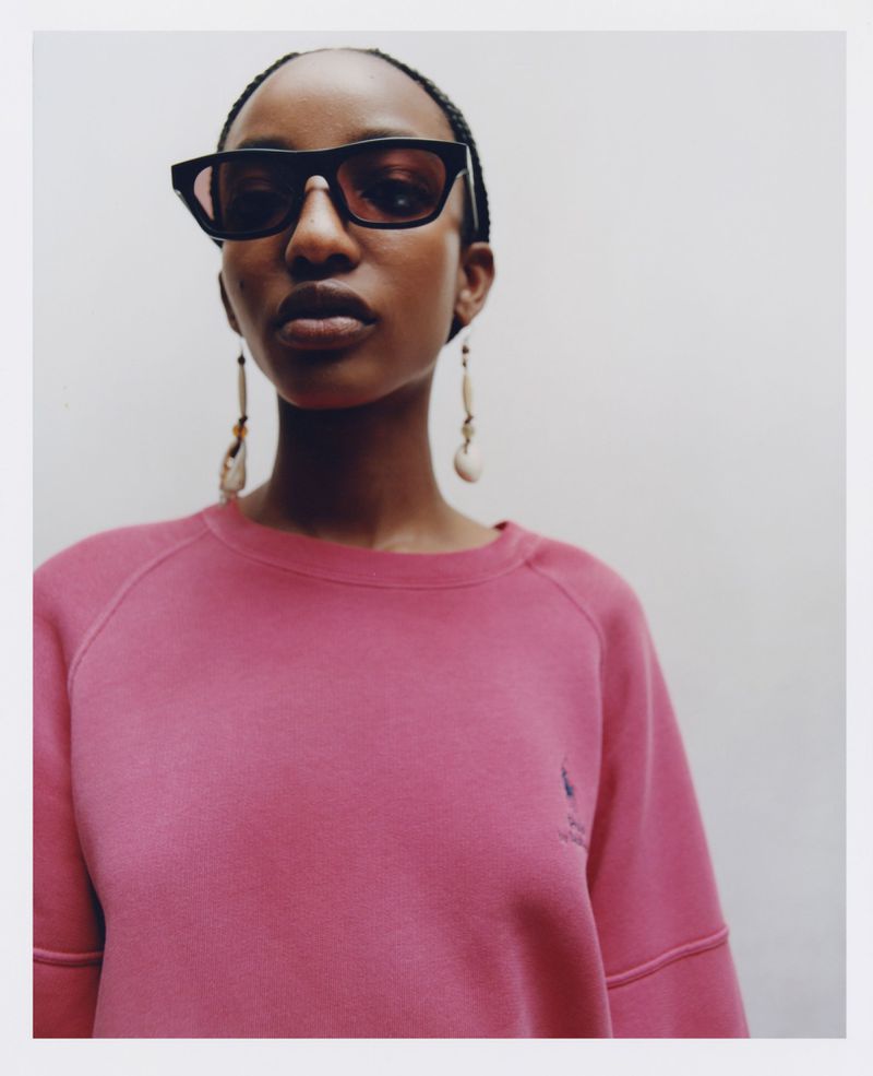 Close-up of an African-American woman wearing sunglasses and a pink sweatshirt. She is centered in the frame against a neutral background.