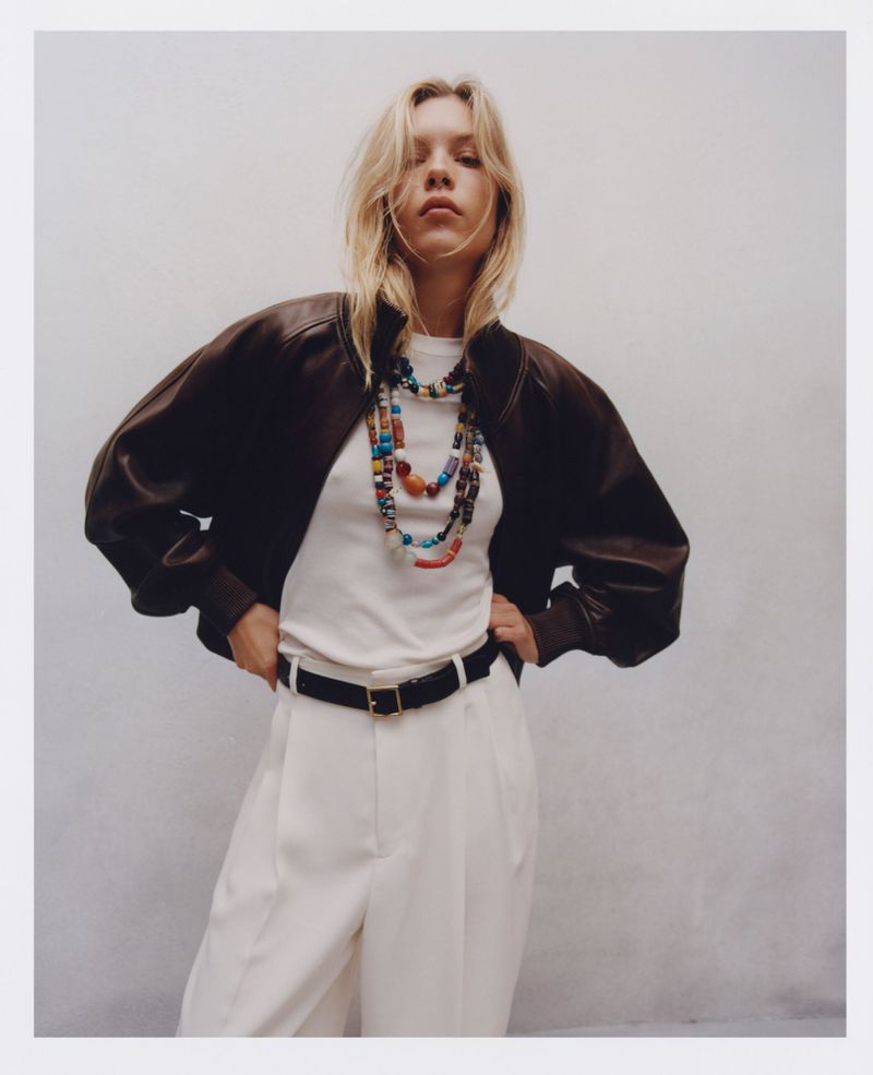 A fashionable woman poses in white pants, a dark jacket, and colorful beaded necklaces against a plain backdrop in a medium studio shot.