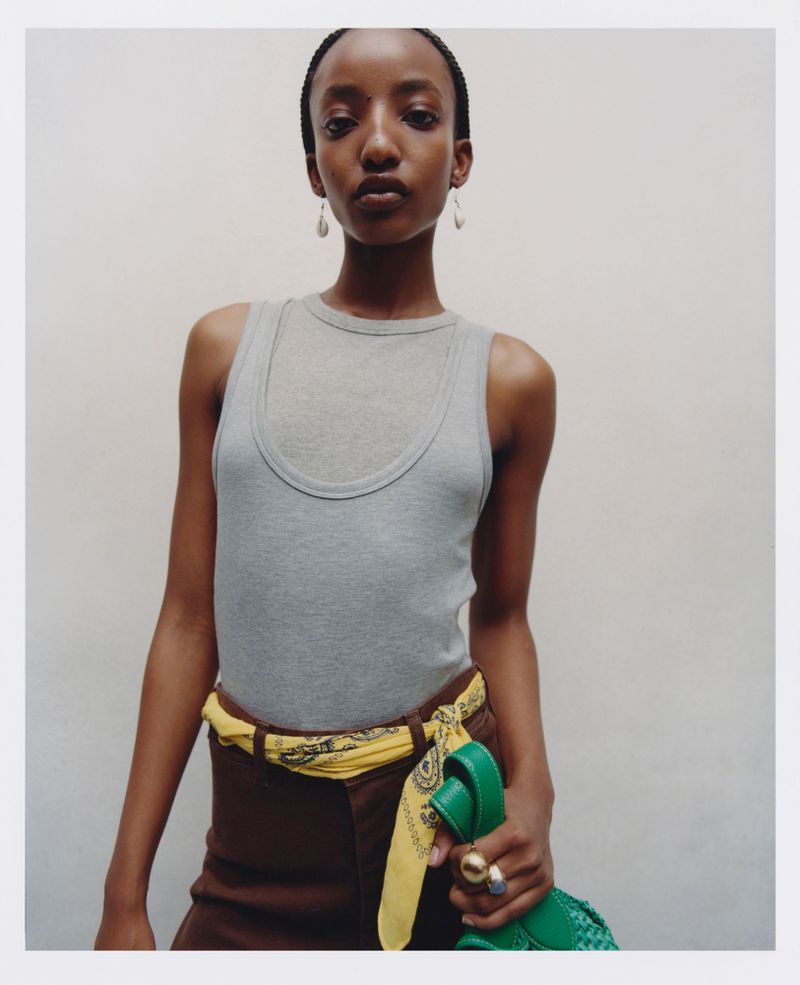 Studio shot of a slender African woman in a grey tank top, brown trousers, and yellow scarf. She is holding a green woven bag.
