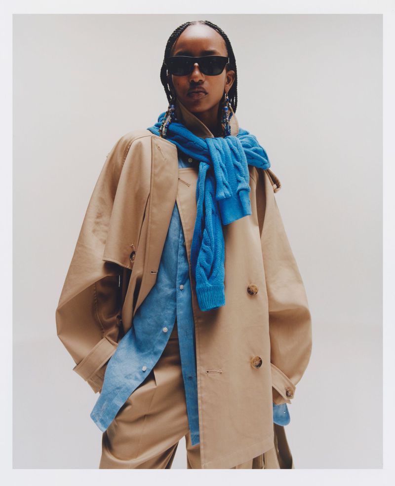 Model wearing dark sunglasses, a tan trench coat, and a draped blue cable-knit sweater against a neutral backdrop.