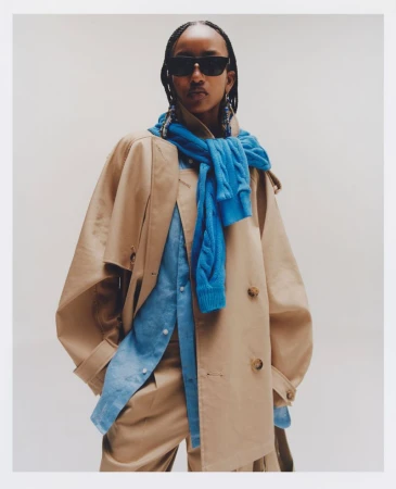 Model wearing dark sunglasses, a tan trench coat, and a draped blue cable-knit sweater against a neutral backdrop.