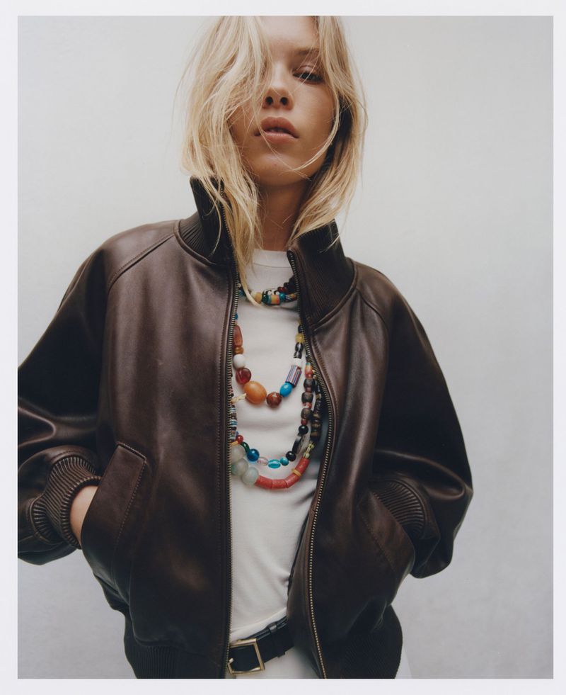 A blonde woman in a brown bomber jacket poses indoors wearing a layered beaded necklace, white top, and dark belt against a neutral wall.