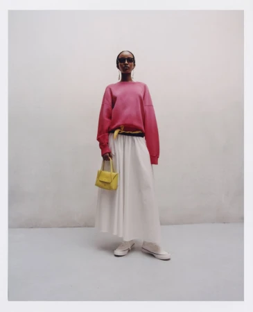 Model Freya Nutter posing in a pink sweatshirt, white maxi skirt, sunglasses, and yellow mini bag in a studio for Polo Ralph Lauren.