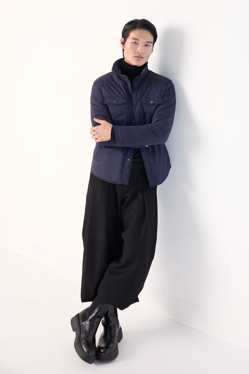 Full-length shot of a slender Asian man posing in a navy quilted jacket, black turtleneck, and wide-legged pants leaning against a white wall.