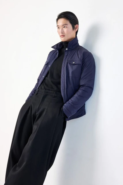 An Asian male fashion model leans against a white wall in a studio, wearing a navy jacket, black turtleneck, and wide-legged black trousers.