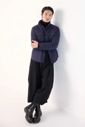 East Asian male model wearing a navy quilted jacket, wide-leg black trousers, and chunky boots leaning against a white studio wall.