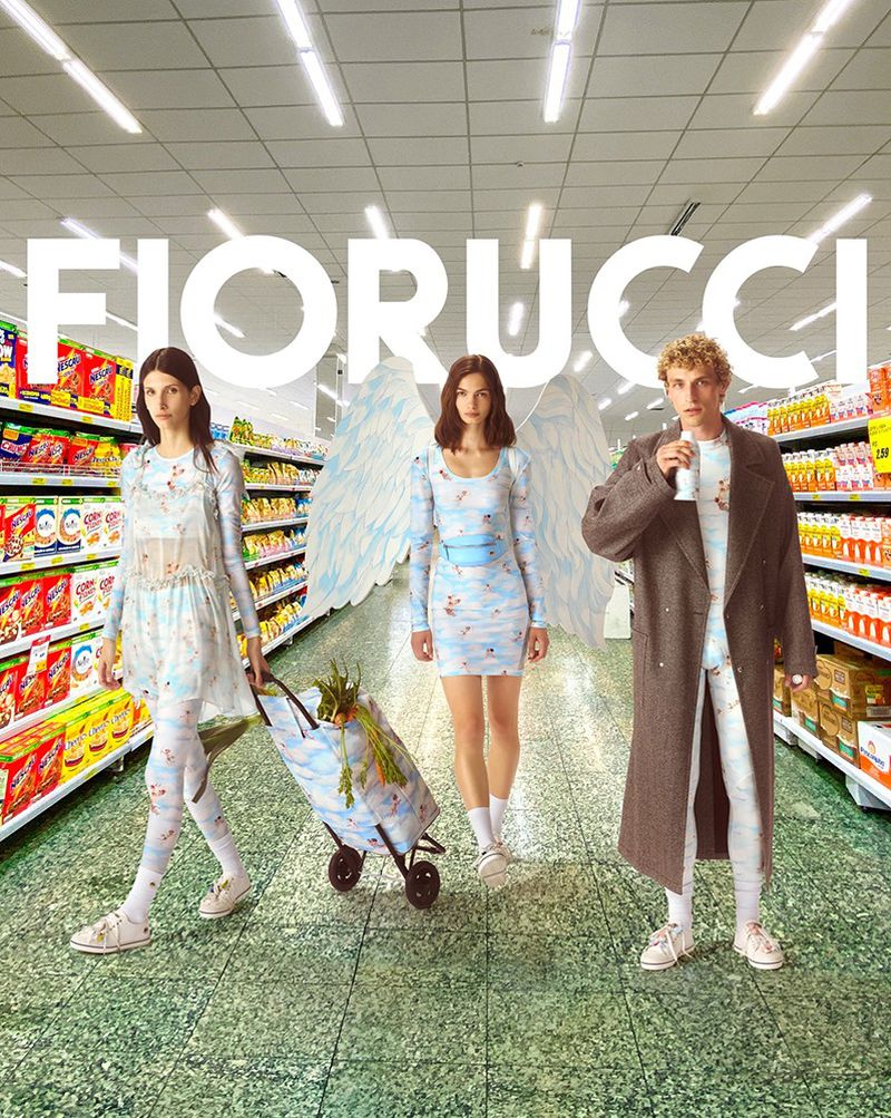 Three fashion models wearing whimsical outfits stand in a grocery store aisle with angel wings. One pulls a shopping cart.