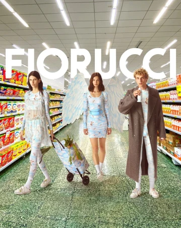 Three fashion models wearing whimsical outfits stand in a grocery store aisle with angel wings. One pulls a shopping cart.