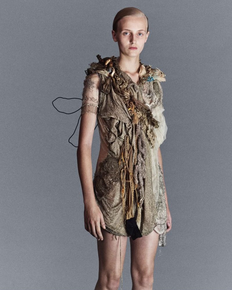 A young woman with short blonde hair poses in a textured tan dress against a gray background in a fashion editorial style photograph.