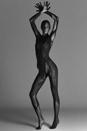 Monochrome image of a slender woman in a patterned bodysuit, hands raised, posing against a smooth gray backdrop in a photo studio.