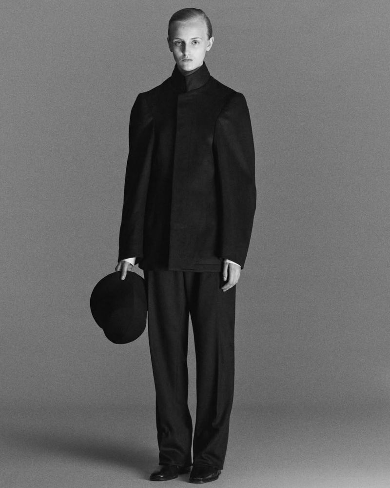 A singular adult female model posing in a full-length shot, wearing a dark, structured suit, and holding a bowler hat in a studio setting.