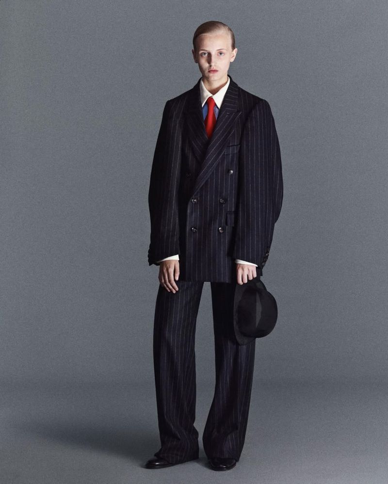 Full-length studio shot of an adult fashion model in an oversized pinstripe suit and black hat against a gray backdrop.