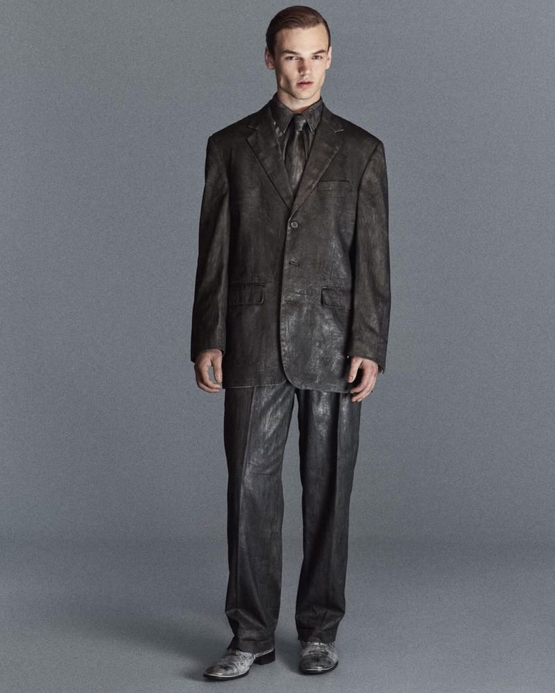 Full-length shot of an adult male model posing in a brown suit and tie against a gray background in a studio setting.
