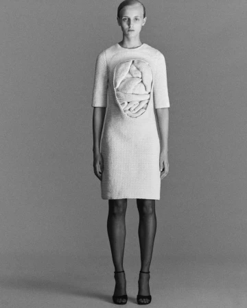 A full shot features a lone woman with an unconventional dress design in a monochromatic studio setting, emphasizing minimalist fashion.