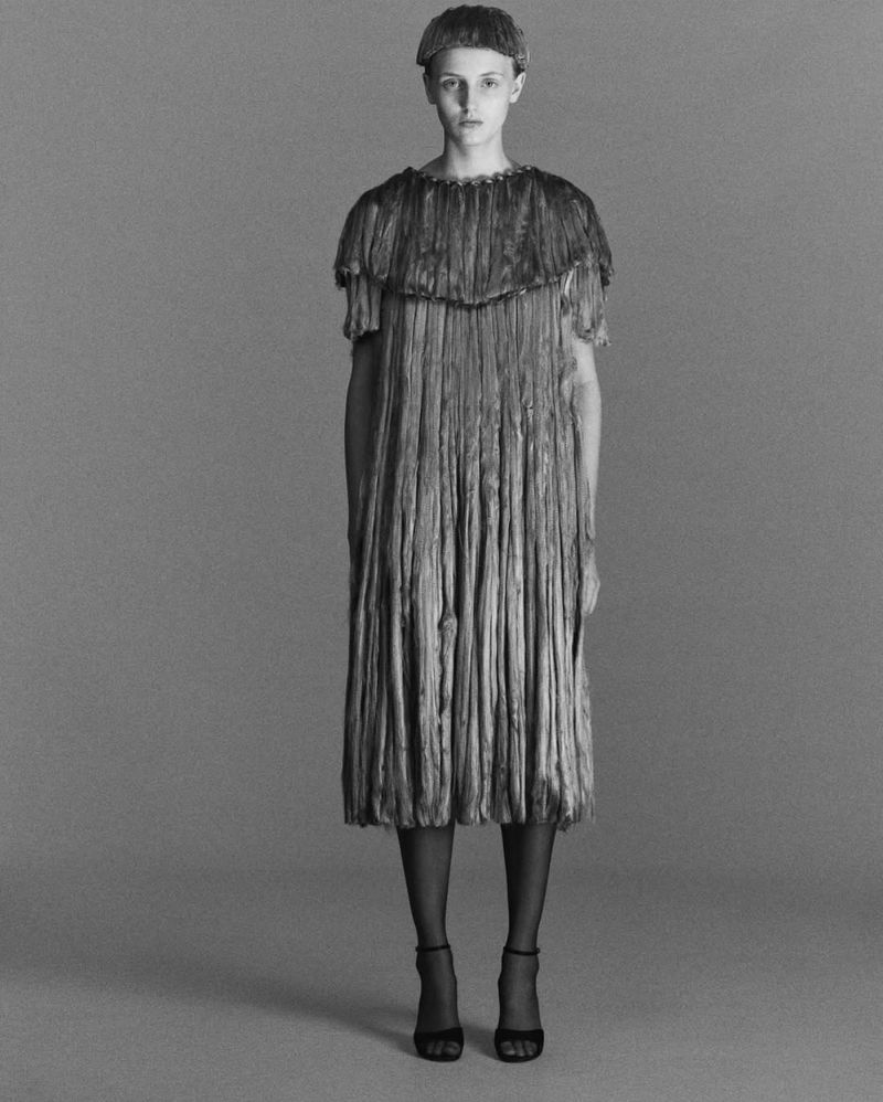 Monochrome portrait of a woman in a layered fringe dress and cap, standing against a grey backdrop. She is wearing high heels.