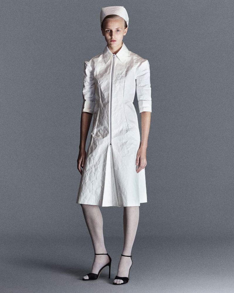 Full-length shot of a model in a white nurse-style dress with zipper, accessorized with a nurse hat and black heels, against a grey backdrop.