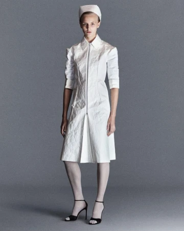 Full-length shot of a model in a white nurse-style dress with zipper, accessorized with a nurse hat and black heels, against a grey backdrop.