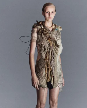 Model wearing a textured deconstructed draped mini dress against a solid grey studio background.