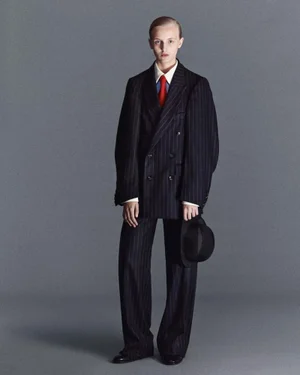 Model wearing an oversized dark pinstripe suit, white collared shirt, and red tie holding a black hat against a grey studio backdrop.