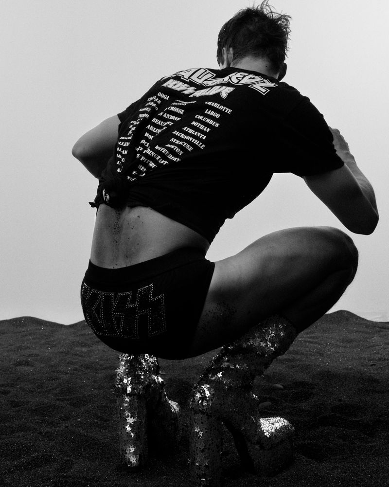 A person in a black shirt and shorts with the word 'KISS' and sparkly boots, crouched on a black sand beach under an overcast sky.
